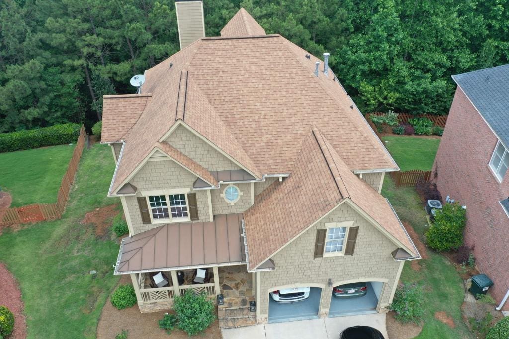 Aerial view of a suburban house with a tan roof, front porch, and two-car garage surrounded by trees